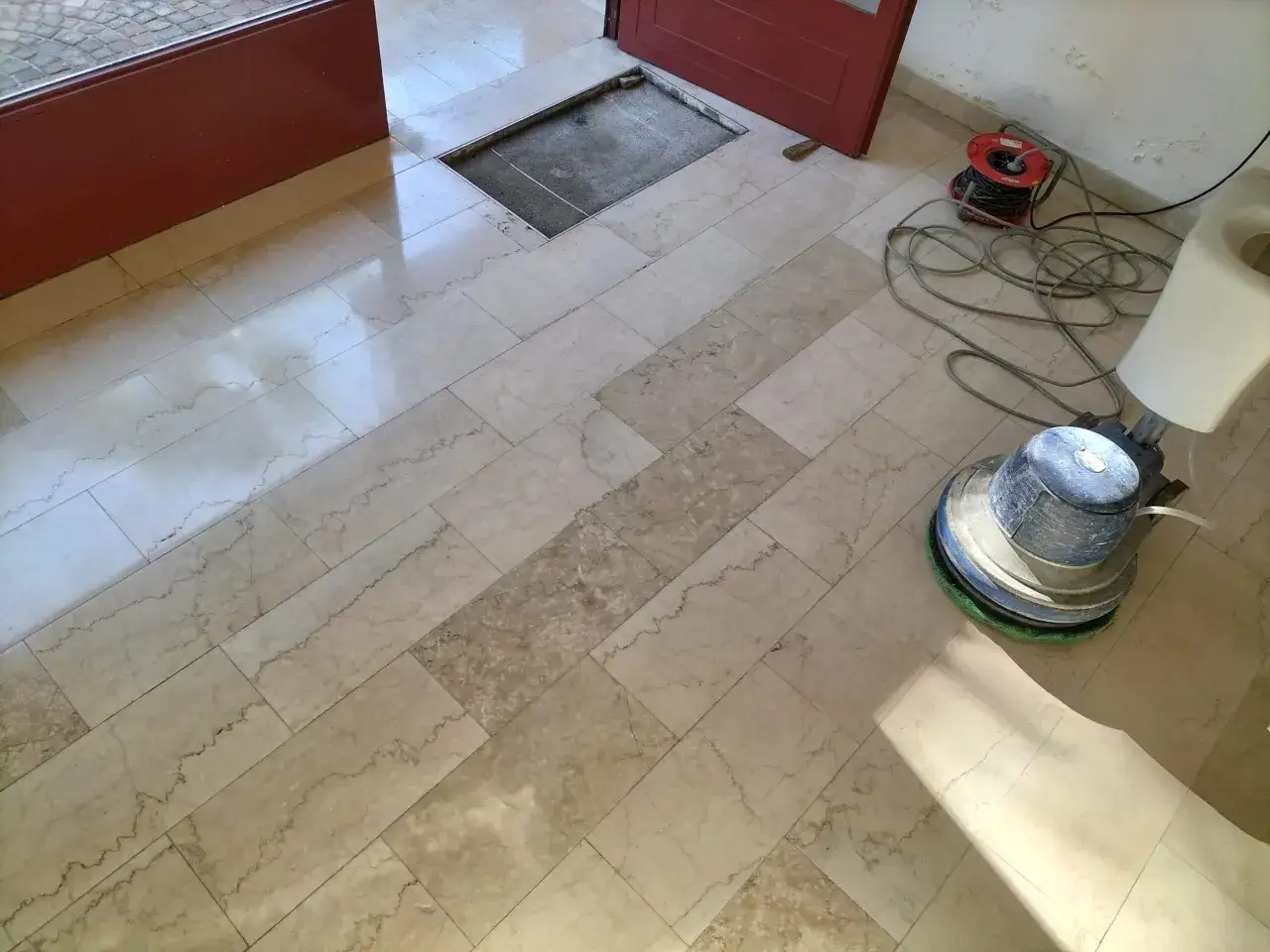 Macchina lucidatrice in azione per mostrare come pulire il marmo vecchio. Pavimento in marmo chiaro con venature scure, cavi elettrici e una porta rossa.