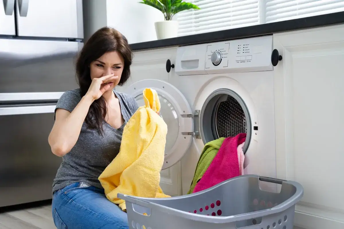 Donna con espressione disgustata, tiene in mano un asciugamano giallo e si tappa il naso. La lavatrice aperta emana un cattivo odore, forse il filtro lavatrice puzza.