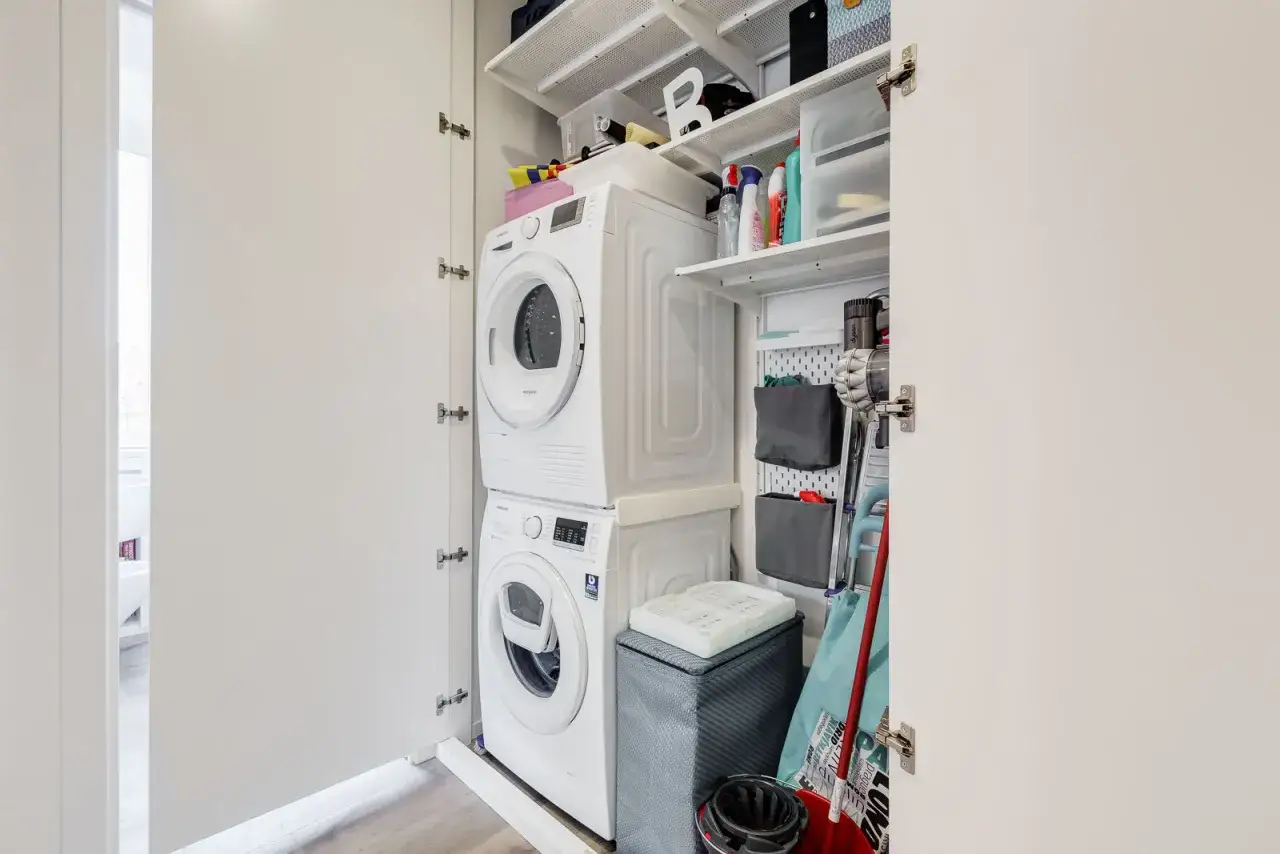 Armadio con lavatrice e asciugatrice impilate. Scaffali pieni di detersivi, contenitori e accessori per la pulizia, un'ottima soluzione per sapere dove mettere i detersivi in bagno.