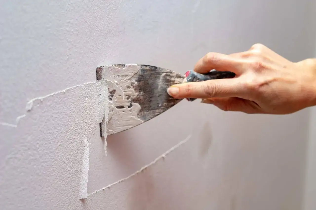 Mano usa una spatola per rimuovere la vernice a smalto dal muro, mostrando il processo di rimozione.