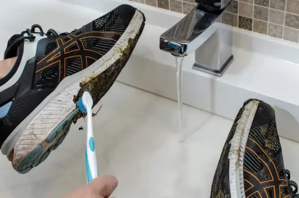 Come lavare le scarpe da running: una mano con uno spazzolino pulisce il fango dalla suola di una scarpa sotto un rubinetto aperto.