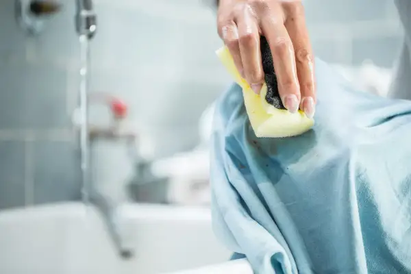 Mano che pulisce una macchia scura su un vestito azzurro con una spugna. Le macchie di profumo vanno via con un po' di pazienza.