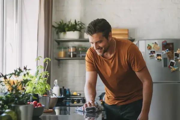 Uomo sorridente pulisce il piano cottura con un panno, mentre sul bancone ci sono piante aromatiche e pomodorini. Forse usa cristalli di soda per lucidare.