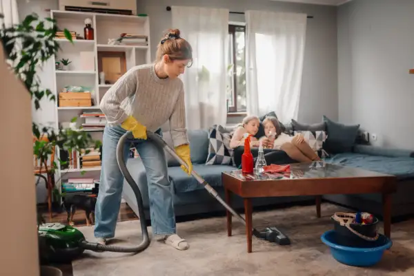 Donna con guanti gialli aspira il tappeto, mentre due bambine guardano il telefono sul divano. Una guida pratica su come pulire una casa sporchissima.